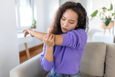 Dirsek ağrısı olan mutsuz bir Asyalı kadın. Ofis sendromu sağlık hizmeti konsepti. Üzgün Asyalı kadının kafası karışmış kollarına bakıyor evinde rahat, parlak bir dairede, koltukta oturuyor..