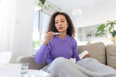 Asyalı kadın grip olmuş ve termometre kullanıyor. Rhinitis hastalığının burnu akıyor. Kanepede uzanmış, termometrede sıcaklığa bakan hasta bir kadın..