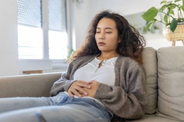 Asian woman lying on sofa looking sick in the living room. Beautiful young woman lying on bed and holding hands on her stomach. Woman having painful stomachache on bed, Menstrual period