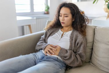 Asian woman lying on sofa looking sick in the living room. Beautiful young woman lying on bed and holding hands on her stomach. Woman having painful stomachache on bed, Menstrual period
