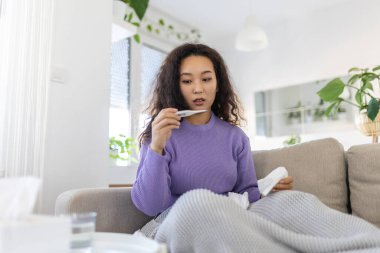 Asian woman is having flu and she is using thermometer. Sick with a rhinitis womanwith hijab has dripping nose. Woman being sick having flu lying on sofa looking at temperature on thermometer.