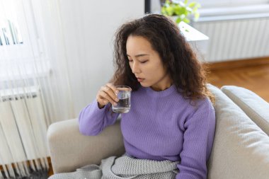 Young Asian woman holding a glass of water at home. Woman on living room sofa Relaxed While Drinking glass of water. Health benefits of drinking enough water