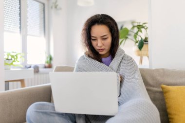 Asian woman wrapped in a cozy blanket sitting on the sofa and working on her laptop