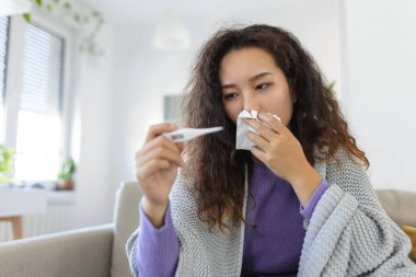 Asian woman is having flu and she is using thermometer. Sick with a rhinitis womanwith hijab has dripping nose. Woman being sick having flu lying on sofa looking at temperature on thermometer.