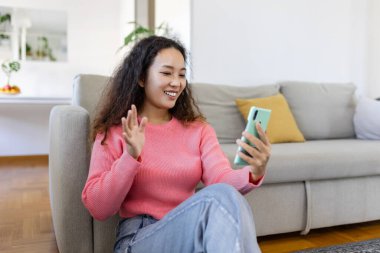 Attractive smiling Asian woman using smart phone while sitting on the sofa at home. Communication and video call concept.