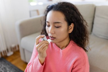 Asian woman using inhaler while suffering from asthma at home. Young woman using asthma inhaler. Close-up of a young Asian woman using asthma inhaler at home.