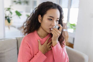 Young Asian woman using her asthma inhaler on couch at home in the living room
