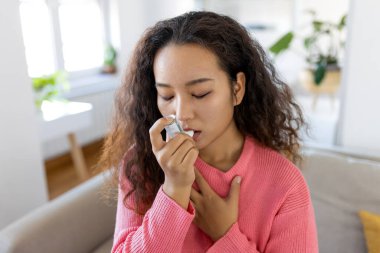 Asian woman using inhaler while suffering from asthma at home. Young woman using asthma inhaler. Close-up of a young Asian woman using asthma inhaler at home.