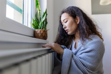 Asian Woman freezing at home, sitting by the cold radiator. Woman with home heating problem feeling cold