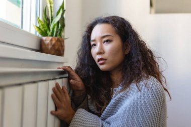 Asian Woman freezing at home, sitting by the cold radiator. Woman with home heating problem feeling cold
