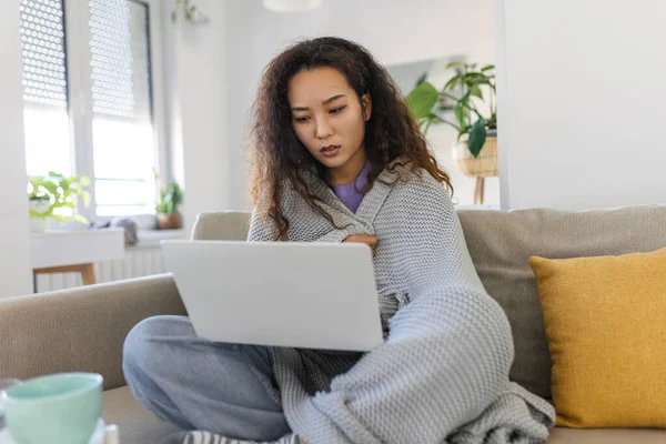 Asian woman wrapped in a cozy blanket sitting on the sofa and working on her laptop