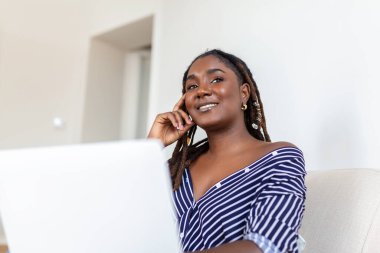 Güzel bir Afrikalı kadın evdeki kanepede dizüstü bilgisayarda çalışıyor. Pazartesi günü benzin dükkanlarında siber satış var mı diye bakıyor. Alternatif ofis için teknoloji kadını konsepti.