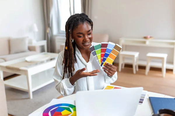 Neşeli Afro-Amerikan bayan tasarımcı müşterilerle video konferansı yapıyor, bilgisayarın önünde oturuyor, elinde renk paletleri tutuyor, el kol hareketi yapıyor ve gülümsüyor.