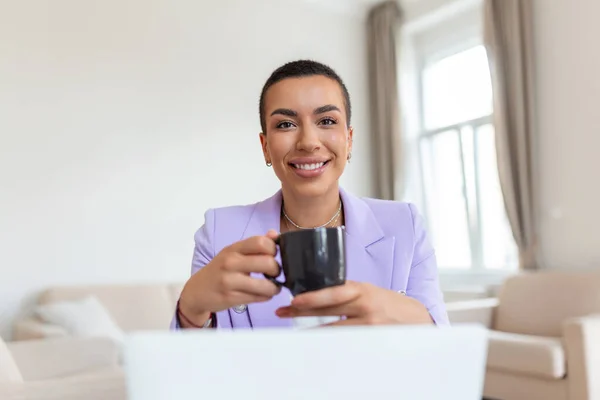 Genç Afrikalı Amerikalı iş kadını uzaktan bilgisayarla çalışıyor ve kahvenin tadını çıkarıyor, gülümsüyor.
