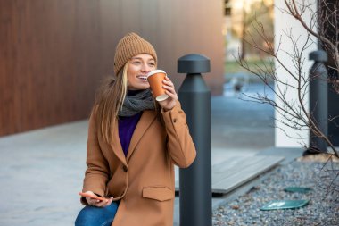 Portrait, Attractive and charming young female in casual clothes sitting on the bench, enjoying her coffee and using her smartphone.