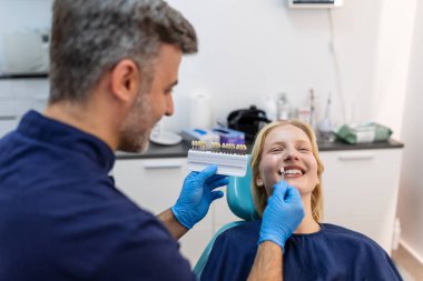 Profesyonel doktor dişlerini düzeltirken dişçi koltuğunda oturan tatmin olmuş genç bir kadının görüntüsü, gülümseyen kadın için dolgu gölgesini seçen dişçi, diş skalası numunesi kullanarak