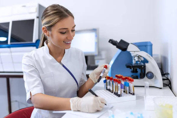 Laboratuarda çalışan kadın bilim adamının yakın plan portresi. Kadın araştırmacı masasında oturmuş mikroskopla kan ve kimyasal test örneklerine bakıyor ve panoya not alıyor. Biyokimya çalışması