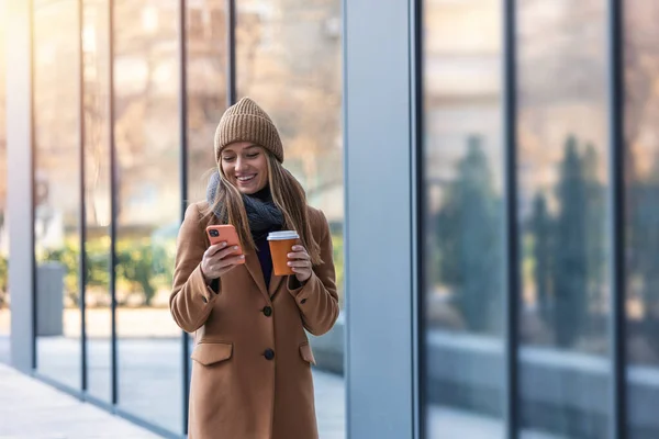 Cheerful young woman wearing coat walking outdoors, holding takeaway coffee cup, using mobile phone