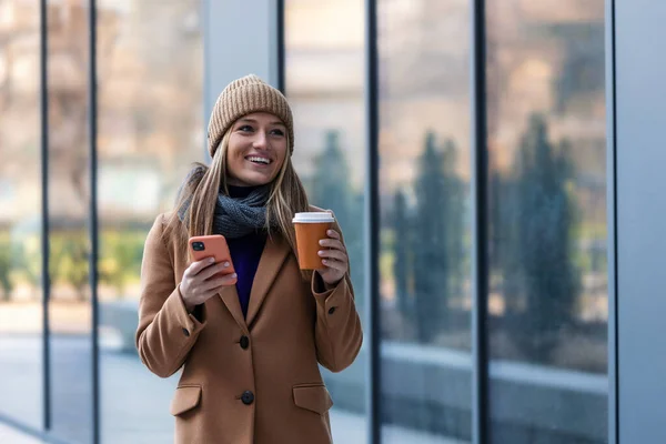 Cheerful young woman wearing coat walking outdoors, holding takeaway coffee cup, using mobile phone