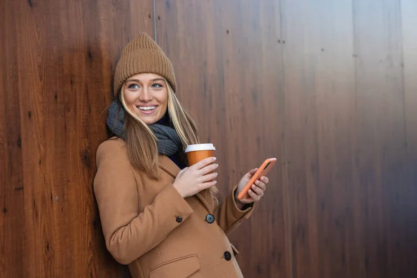 Cheerful young woman wearing coat using her phone in the sunny city street and drinking take away coffee in paper cup.
