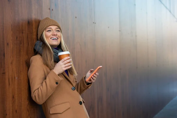 Cheerful young woman wearing coat using her phone in the sunny city street and drinking take away coffee in paper cup.