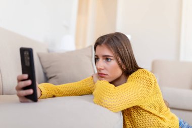 Dalgın, üzgün, genç bir kadın elinde akıllı telefonuyla erkek arkadaşından mesaj bekliyor..