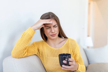 Evde kanepede kahve içerken baş ağrısı olan depresif genç kadın.