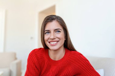 Evdeki kanepede oturan ve kameraya bakan mutlu genç kadın. Kışlık giysiler içinde rahat bir kadın portresi koltukta dinleniyor. Güz boyunca gülümseyen ve rahatlatan güzel bir kadının portresi..