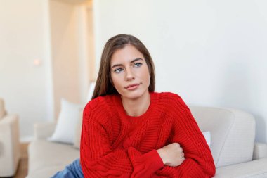 Evdeki kanepede oturan ve kameraya bakan mutlu genç kadın. Kışlık giysiler içinde rahat bir kadın portresi koltukta dinleniyor. Güz boyunca gülümseyen ve rahatlatan güzel bir kadının portresi..