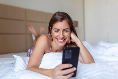 Beyaz yatakta yatarken cep telefonuna bakan gülümseyen kadın. Evde cep telefonu kullanan mutlu esmer genç bayan. Akıllı telefondan internette sörf yapan güzel bir kız.
