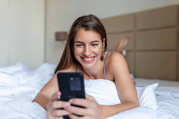 Beyaz yatakta yatarken cep telefonuna bakan gülümseyen kadın. Evde cep telefonu kullanan mutlu esmer genç bayan. Akıllı telefondan internette sörf yapan güzel bir kız.