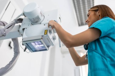 Doktor klinikteki röntgen cihazına bakıyor. Kadın doktor makineyi hastanın röntgenini çekmek için ayarladı. Radyolog ve hasta röntgen odasında..