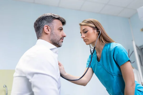 Genç doktor steteskop kullanıyor. Hastanın kalp atışlarını dinle. Kadın bir doktorun erkek bir hastaya muayene yaparken görüntüsü.