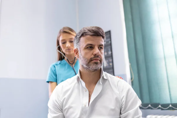 Klinikteki kadın bir doktor, koronavirüs veya zatürree belirtileri gösteren bir hastanın akciğerlerini kontrol eder. Doktor öksürüyor. Ciğerlerindeki hırıltıyı duyuyor.