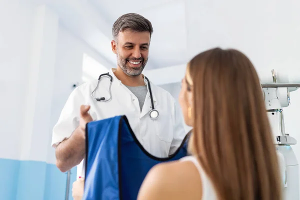 Klinikteki X-ray muayene hastası. Doktor X-ray 'in önündeki hastaya mavi koruyucu giysi veriyor. Erkek Radyolog, X-ray Bilgisayar Tomografisi için Kadın Hastayı Hazırlıyor