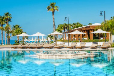 Limasol, Kıbrıs - 19 Mayıs 2021: Lüks modern otelin havuz alanı, palmiye ağaçları ve tropikal bitkilerin gölgesinde güneşli yataklar