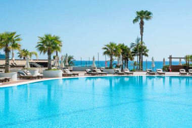 Limasol, Kıbrıs - 19 Mayıs 2021: Lüks oteldeki havuz alanı, palmiye ağaçları ve tropikal bitkilerin gölgesinde güneşli yataklar