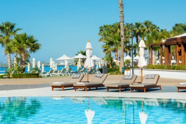 Limasol, Kıbrıs - 19 Mayıs 2021: Lüks modern otelin havuz alanı, palmiye ağaçları ve tropikal bitkilerin gölgesinde güneşli yataklar