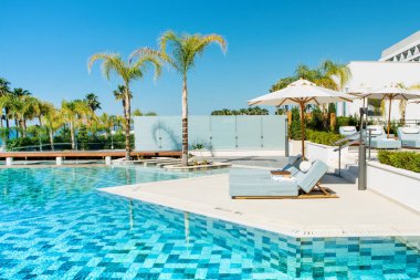 Limasol, Kıbrıs - 19 Mayıs 2021: Lüks modern otelin havuz alanı, palmiye ağaçları ve tropikal bitkilerin gölgesinde güneşli yataklar