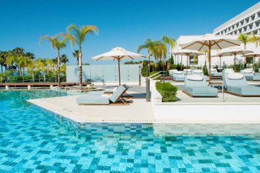 Limasol, Kıbrıs - 19 Mayıs 2021: Lüks oteldeki havuz alanı, palmiye ağaçları ve tropikal bitkilerin gölgesinde güneşli yataklar