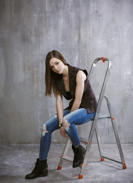 Молодая, стройная девушка сидит на стремянке на фоне серого — стоковое фото