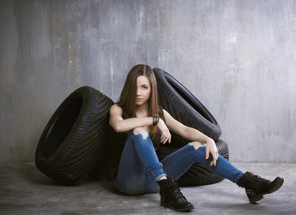 Joven, chica delgada sentado en neumáticos de coche en un fondo de gris co — Foto de Stock