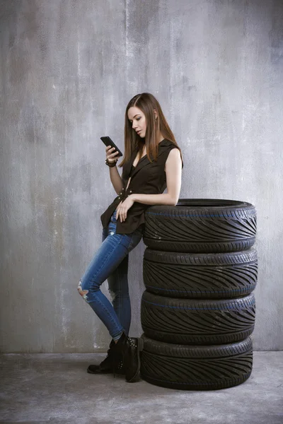 Chica joven con un teléfono móvil, se apoya contra los neumáticos en — Foto de Stock