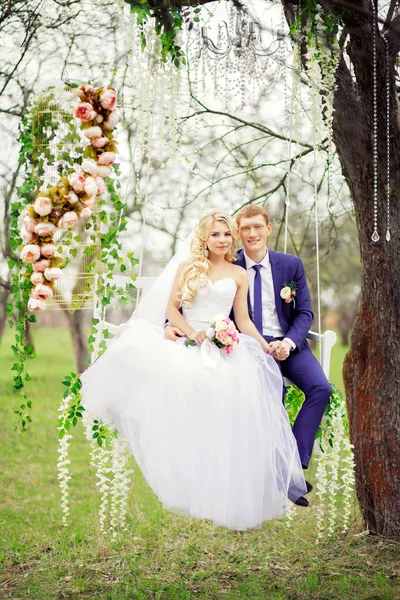 Junge und schöne Braut und Bräutigam sitzt auf einer weißen Schaukel in — Stockfoto