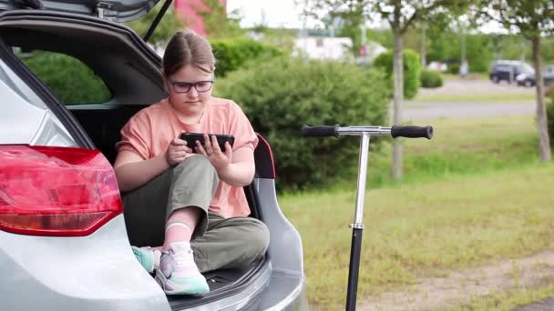 Linda niña preadolescente sentada en el coche, usando un teléfono inteligente ignorando su scooter en el fondo. Niños usando el concepto de tecnología. — Vídeo de stock