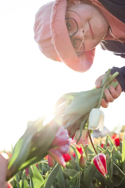 Jovem adolescente usando óculos, segurando um grande monte de tulipas e espreitando para a câmera. — Fotografia de Stock