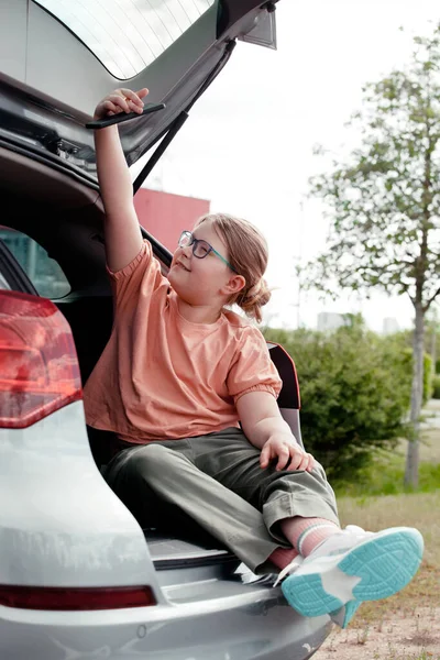 Linda niña preadolescente sentada en el coche, usando un teléfono inteligente ignorando su scooter en el fondo. Niños usando el concepto de tecnología. Imagen De Stock