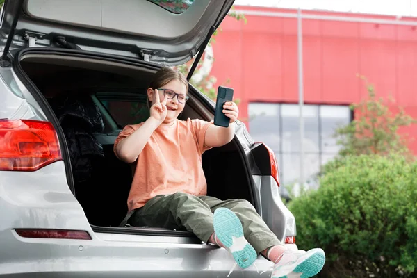 Nettes preteen Mädchen sitzt im Auto und verwendet Smartphone ignoriert ihren Roller auf dem Hintergrund. Kinder nutzen Technik-Konzept. lizenzfreie Stockfotos