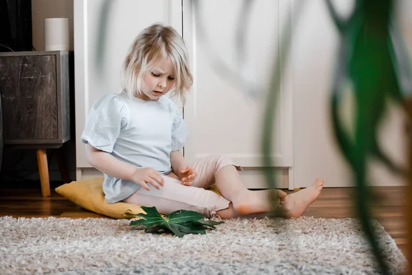 Petite fille assise sur le sol dans le salon et jouant avec des feuilles de plantes exotiques — Photo