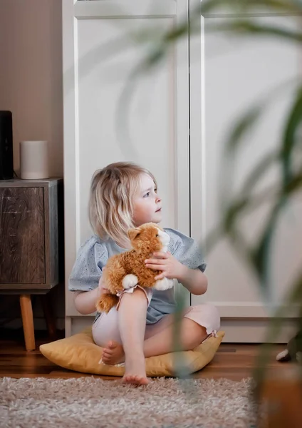 Mała dziewczynka siedząca na podłodze w salonie i bawiąca się kotem zabawki, strzał wewnątrz, naturalne światło — Zdjęcie stockowe